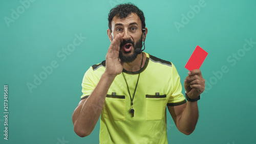 Photography Man referee in neon shirt holds red card and whistle, hand cupped to mouth as if shouting, headset mic and beard visible in studio setting; discipline stern