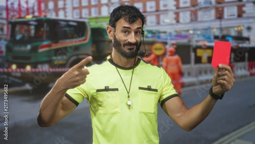 Quadro su tela Man referee holding red card and pointing index finger on street near construction lane with truck and workers; firm authority
