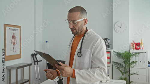 Konstfotografi Man doctor with stethoscope and clipboard reads and writes notes in clinic building beside crutches, anatomy poster and wall clock; focused care