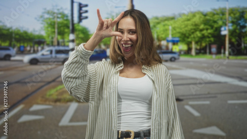 Quadro su tela Woman making i love you hand sign with bare hand and smiling at a city street intersection with cars and traffic light; joy travel