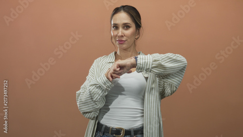 Fotografía Woman points finger to wristwatch while checking time, casual striped shirt and white tank top visible in studio; impatience punctuality waiting