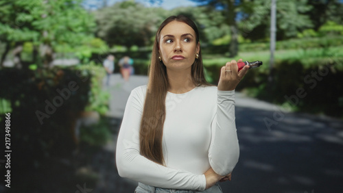 Young woman holding a vape with fingers near her mouth in a brief inhale gesture on a street path with blurred pedestrians and trees behind; contemplation calm.