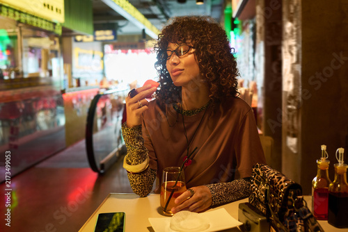 Young adult biracial woman with curly hair enjoying food in cafe with eyes closed. Image represents leisure and dining out, suitable for food blogs and lifestyle marketing