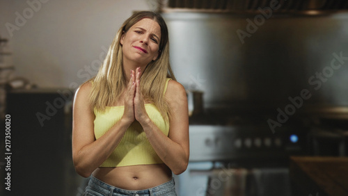 Foto Young woman with palms together and pleading face in a commercial kitchen wearing a yellow tanktop and jeans; pleading forgiveness