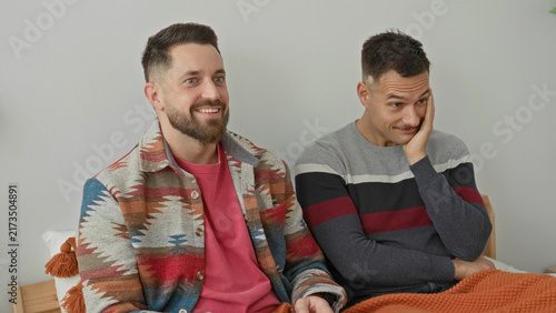 Obraz na płótnie Two men sitting on a bed, one pointing a tv remote while smiling and the other rests hand on cheek in a studio; playful intimacy