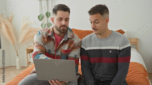 Two men sit on a bed  one holds a laptop while both smile and exchange a highfive in a cozy bedroom with orange blanket and pampas grass  planning intimacy joy.