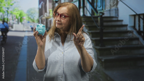 фотографія Senior hispanic woman holds blue piggy bank and points finger toward viewer on a street beside building steps; savings caution