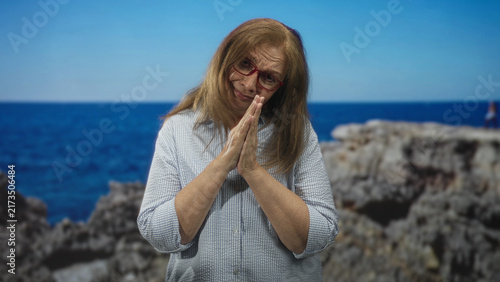 Obraz na plátně Woman with hands pressed together in prayer pose at studio seaside backdrop wearing red glasses and striped shirt; hopeful reflection