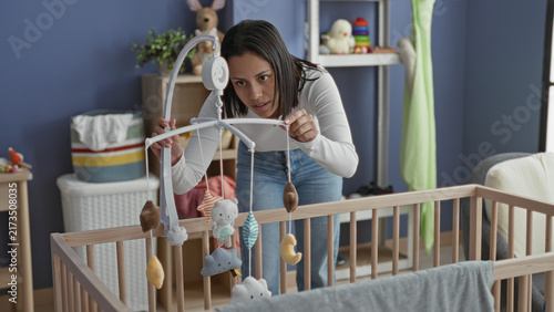 Fotografía Woman adjusts a hanging baby mobile over a wooden crib, points finger to a plush toy and leans in amid nursery furniture in building; tenderness