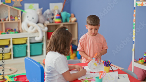 Bild auf Leinwand Boy holds orange scissors and girl draws on paper at a building playroom table with colored pencils and crafts; playful friendship