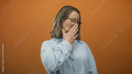 Woman grey hair senior covers mouth with hand in a studio with orange backdrop, wearing glasses, light blue shirt and gold necklace; disgust discomfort.