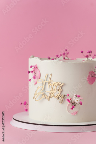 Elegant white birthday cake with pink blossoms and gold Happy Birthday topper on the pink background