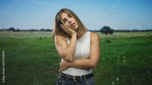 Obraz na plátně Woman hand on cheek, tilted head and crossed arms, wearing white tank and jeans in a forest meadow outdoors, daylight portrait; thoughtful reflection
