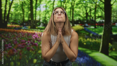 Quadro su tela Woman pressing hands together in prayer in a flower filled park, glasses, sleeveless top, looking upward; hopeful reflection