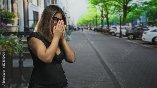 фотографія Woman covers mouth with hands and wears glasses while standing on a tree lined street near parked cars; surprise privacy vulnerability