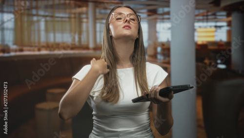 Obraz na płótnie Woman holding tablet touching her forehead in an airport lounge, glasses on and eyes closed, mid exhale and sigh; relief pause