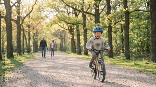 Young boy rides bicycle on park path with parents walking behind