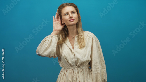 Bild auf Leinwand Woman cupping hand to ear in studio against blue backdrop wearing beige dress and necklace; curiosity attentiveness