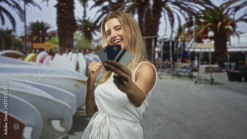 фотографія Woman holding creditcard and raising fist in celebratory pose on street near surfboards and palm trees; shopping joy vacation