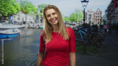 Woman smiling at camera, standing by a canal with boat and bicycle on a street lined with lampposts and buildings; cheerful warmth.