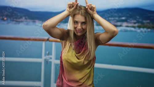 Принти на полотні Blonde woman on ship deck making horn gesture with fingers, bare forearms raised over head, cruise railing and sea visible behind her; playful vacation