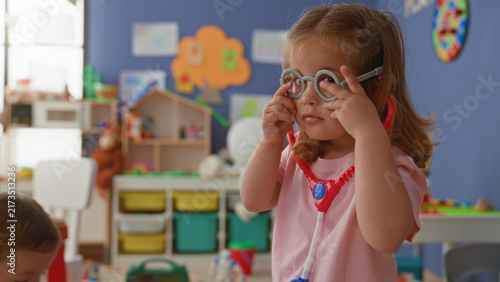 Obraz na plátně Girl covers eyes with hands while wearing toy stethoscope and play glasses in preschool building; curiosity imaginative play