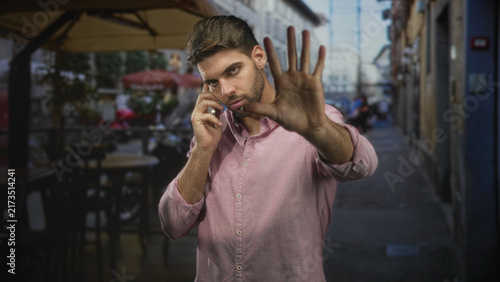 Принти на полотні Man with beard holding smartphone to ear, palm raised in stop gesture toward camera, standing at a street cafe sidewalk with tables and umbrellas; annoyance privacy