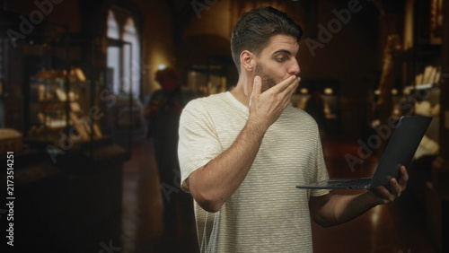 Obraz na płótnie Man with beard holds laptop and touches chin while gesturing with free hand in a museum building, studying the screen intently; curiosity