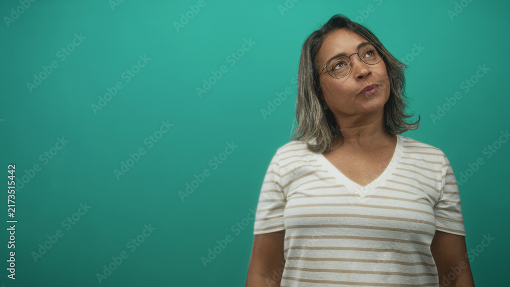 Fototapeta premium Middle aged hispanic woman in a striped v neck shirt wearing glasses, head tilted and eyes closed against a teal studio backdrop; quiet contemplation.