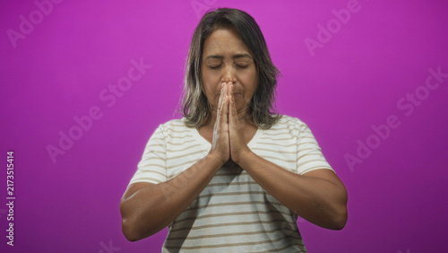 Fotografie Hispanic woman with hands pressed together in prayer gesture in studio against magenta backdrop, eyes closed and striped shirt; serenity gratitude reflection
