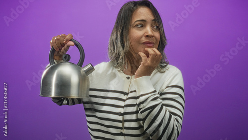 Fotografía Woman in striped sweater holds stainless kettle by handle and touches chin with hesitant expression in studio; quiet contemplation
