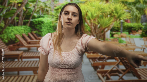 Fotografie Woman hispanic pointing arm toward lounge chair and pool deck in building wearing pink dress and neutral expression; determination