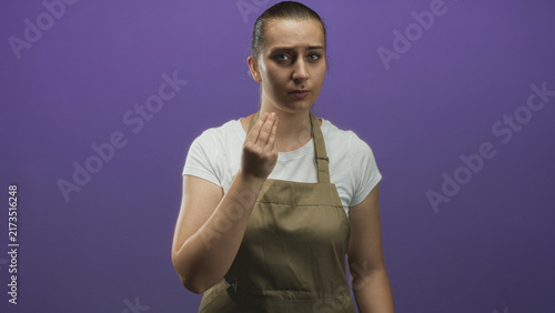 Картината върху платно Woman wearing a khaki apron beckons with hand and furrows brow toward camera in studio setting; impatience service