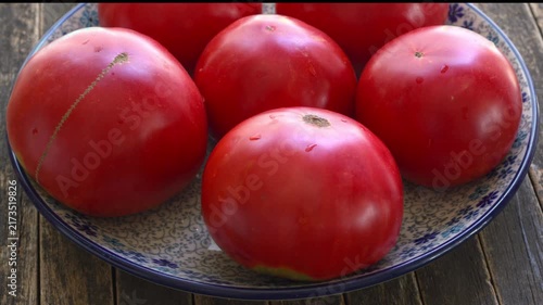 Brandywine heirloom tomatoes rotating on a floral ceramic plate. Large organic heritage tomatoes on a dark, rustic wood-plank background, slowly spinning in a tilted view.