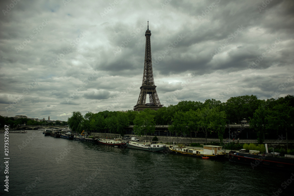 Fototapeta premium A Wide Scenic View of a Famous Iron Lattice Tower next to a River with Moored Boats Under a Cloudy Sky