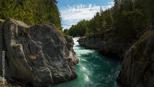 Fotografie An aerial wide low shot gets a closer perspective inside the rocky canyon walls as the turquoise Cheakamus River flows onward from the distant rushing waterfalls under a blue sky