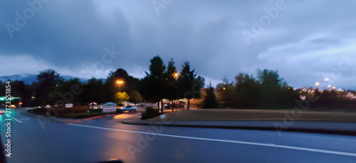 Soft focus long exposure shot from a moving car window of a wet asphalt road and shopping center behind trees at twilight with blurry streetlights reflecting on rainy pavement

