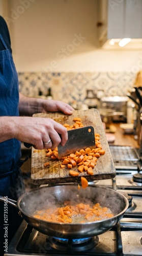 Close-up of hands scraping chopped vegetables from a wooden cutting board into a hot stainless steel pan