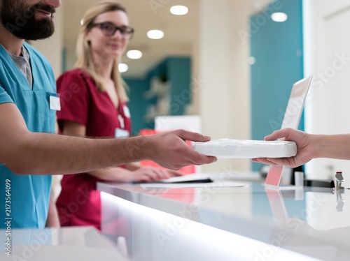 A healthcare professional hands over a package to a colleague in a modern clinic. The two are wearing scrubs and appear to be in a medical setting, possibly a reception or ward area.