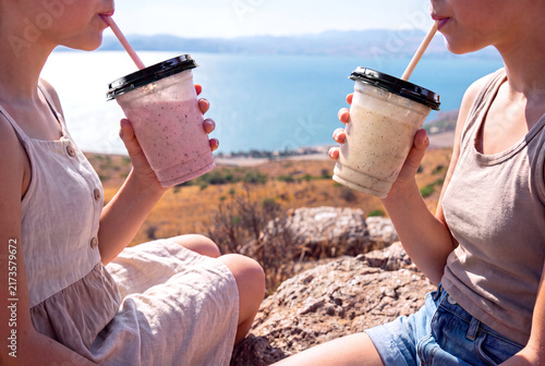 Close view of two women drinking pale fruit and milk smoothies from plastic cups with straws, with a softly blurred blue sea, distant coastline, cliffs and sunlit landscape behind.