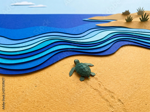 A small sea turtle walking on the sandy beach towards the blue paper craft ocean waves.
