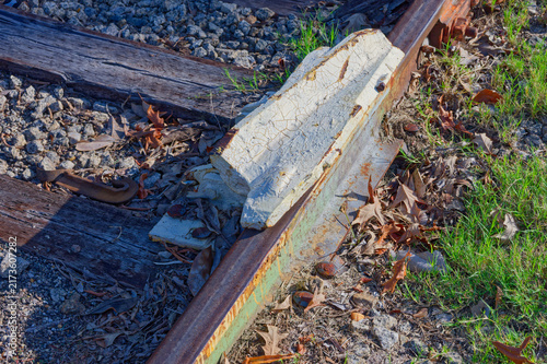 Fotografie Rusty railroad track and weathered wooden ties surround a cracked concrete rail anchor amid gravel, grass and fallen leaves, evoking texture, decay and industrial nostalgia in natural light