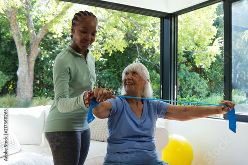 Фотография Senior woman on sofa using blue band while African American female trainer guidi