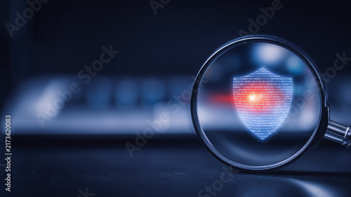A magnifying glass focused on a digital shield represents security inspection cyber protection and technology risk assessment.
