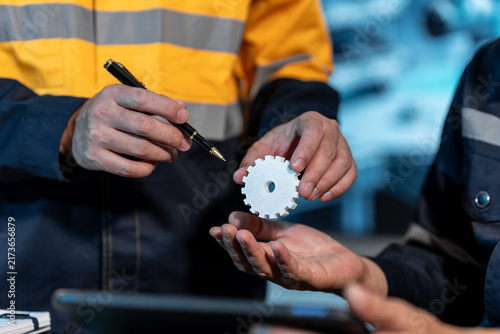 Close-up of industrial workers inspecting a small white gear in a factory, showing teamwork, equipment maintenance and quality control.