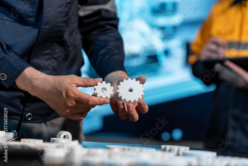 Close-up of a technician holding two small white plastic gears in a workshop, with tools, machine parts, and a coworker blurred in the background.