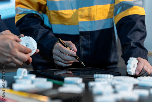 Two industrial workers inspect plastic gears and record measurements on a tablet at a factory workbench.