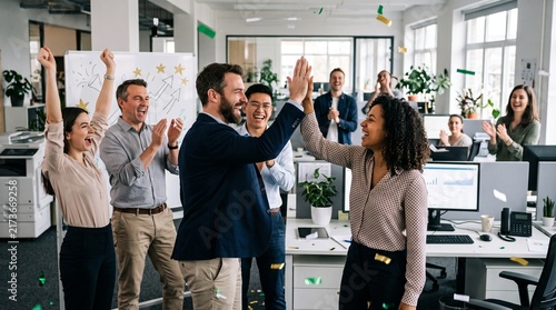 Celebrating Success: A group of smiling business colleagues high-fives in an office setting, surrounded by a celebratory atmosphere of achievement and recognition.