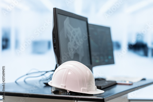 Fotografie White safety helmet on a desk beside dual computer monitors in a bright modern office, representing engineering, construction planning, workplace safety and design work