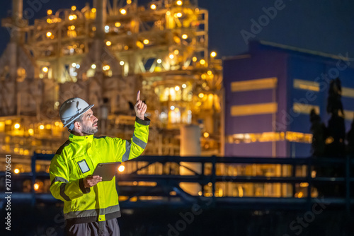 Inspector worker inspecting petroleum engineering in oil refinery performance checklist inspection at fuel industrial plant Male engineer pointing up and using tablet at site during night time